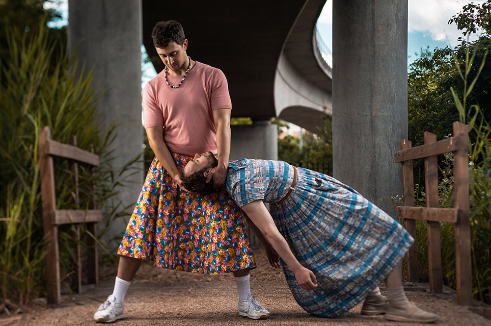 Zwei Schauspieler in farbigen Frauenkleidern in akrobatischer Pose vor urbanem Hintergrund.