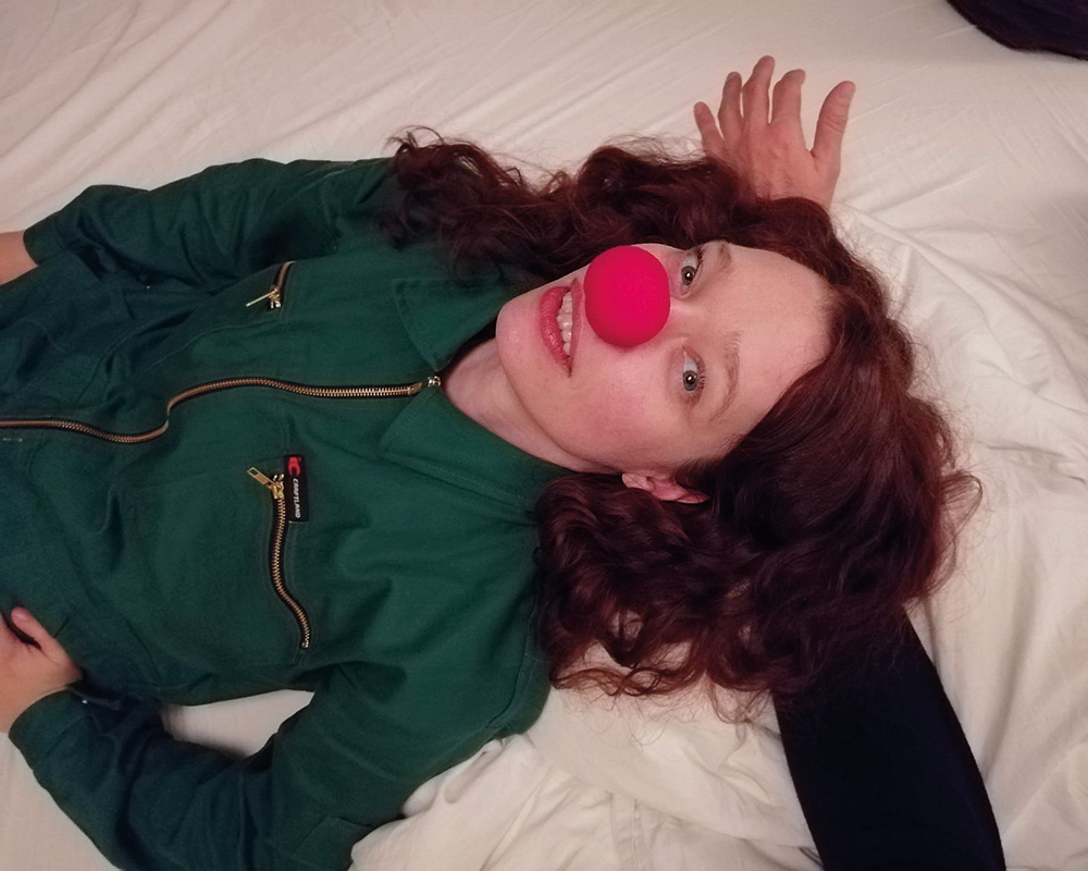Schauspielerin liegt auf einem Bett und hat eine Clownsnase auf.