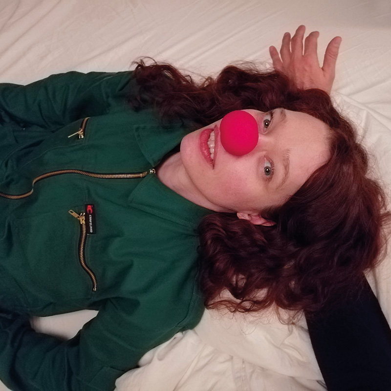 Schauspielerin liegt auf einem Bett und hat eine Clownsnase auf.
